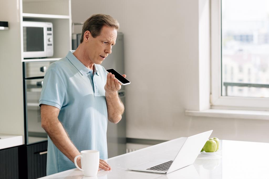 foco seletivo do homem gravando mensagem de voz enquanto mantém o smartphone com tela preta perto do laptop
  - Foto, Imagem
