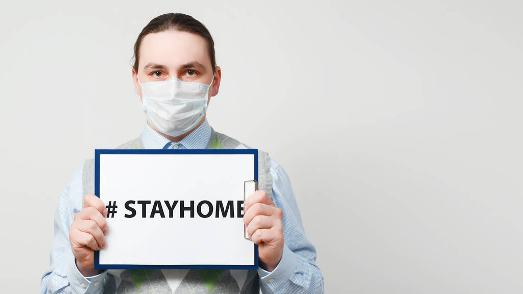 Άντρας γιατρός με προστατευτική μάσκα κρατά χαρτί με το hashtag μείνετε στο σπίτι σε γκρι φόντο. - Φωτογραφία, εικόνα