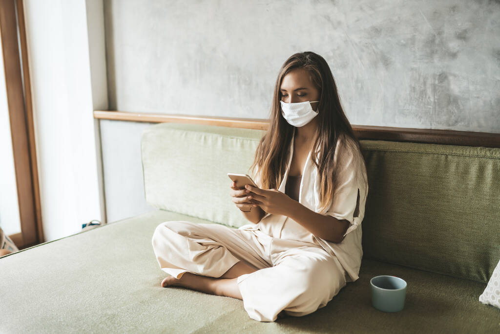 Mujer joven con máscara médica usando el teléfono sentado en el sofá en casa
 - Foto, imagen