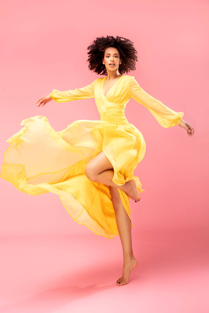 Afro mladá žena tanec ve žluté maxi šaty přes růžové pastelové studio pozadí. - Fotografie, Obrázek