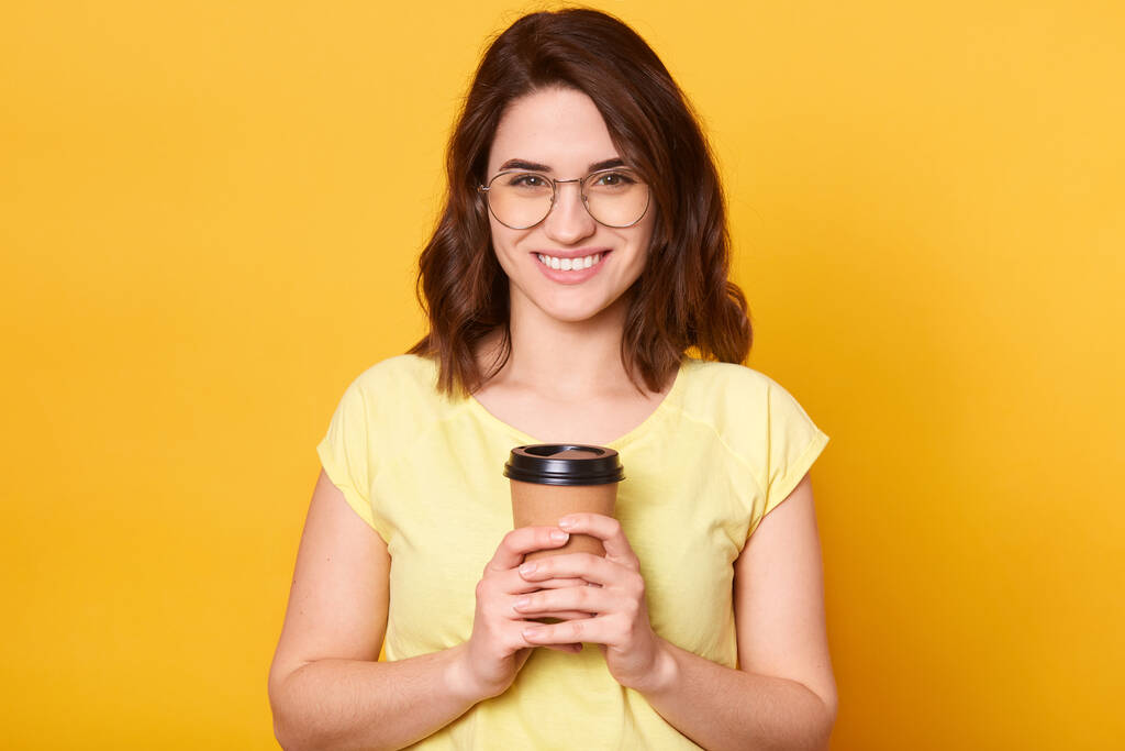 Horizontal shot of smiling happy brunette female wearing casual t shirt, holding takeaway coffee in both hands, looking at camera with smile, enjoying hot beverage, winsome girl with charming smile. - Photo, Image