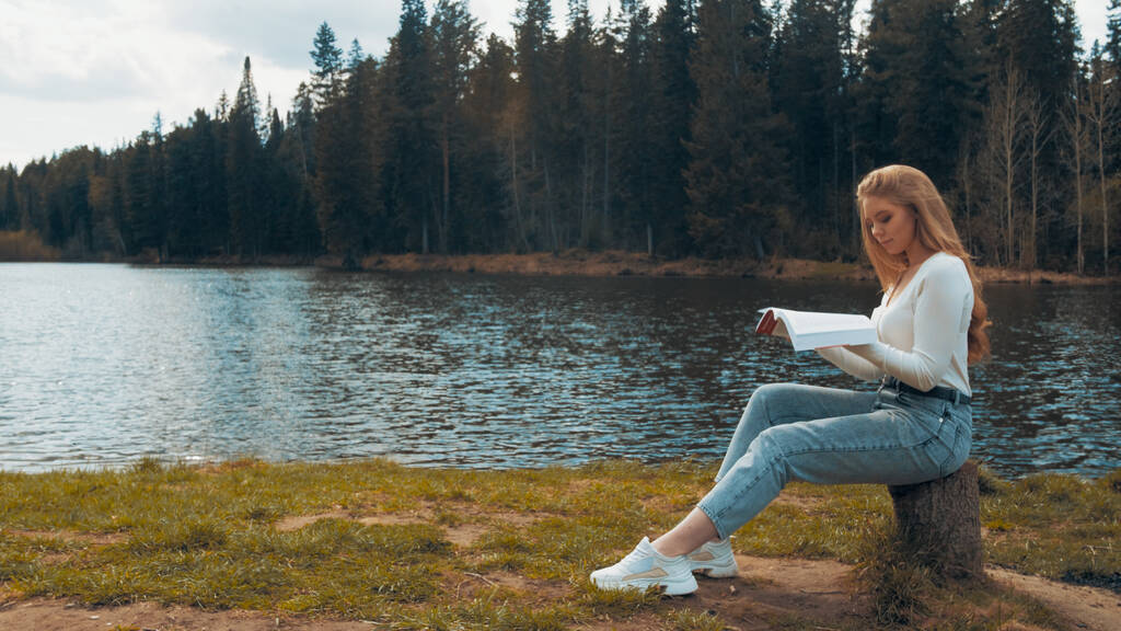 Mooie jonge blonde vrouw zit en leest boek in een park in de buurt van het meer. Zijaanzicht - Foto, afbeelding