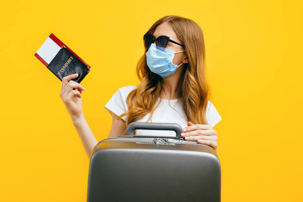 Μια γυναίκα ταξιδιώτης με μια ιατρική προστατευτική μάσκα στο πρόσωπό της, κρατώντας εισιτήρια και διαβατήριο, στέκεται με μια βαλίτσα σε κίτρινο φόντο. Έννοια της καραντίνας, coronavirus, ταξίδια - Φωτογραφία, εικόνα