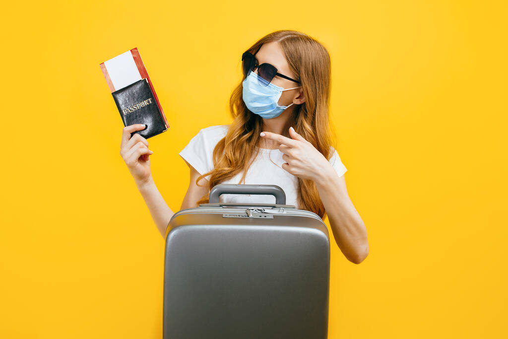 Yüzünde tıbbi koruyucu maske takan, elinde biletler ve pasaportla sarı arka planda duran bir kadın yolcu. Karantina, koronavirüs, seyahat kavramı - Fotoğraf, Görsel