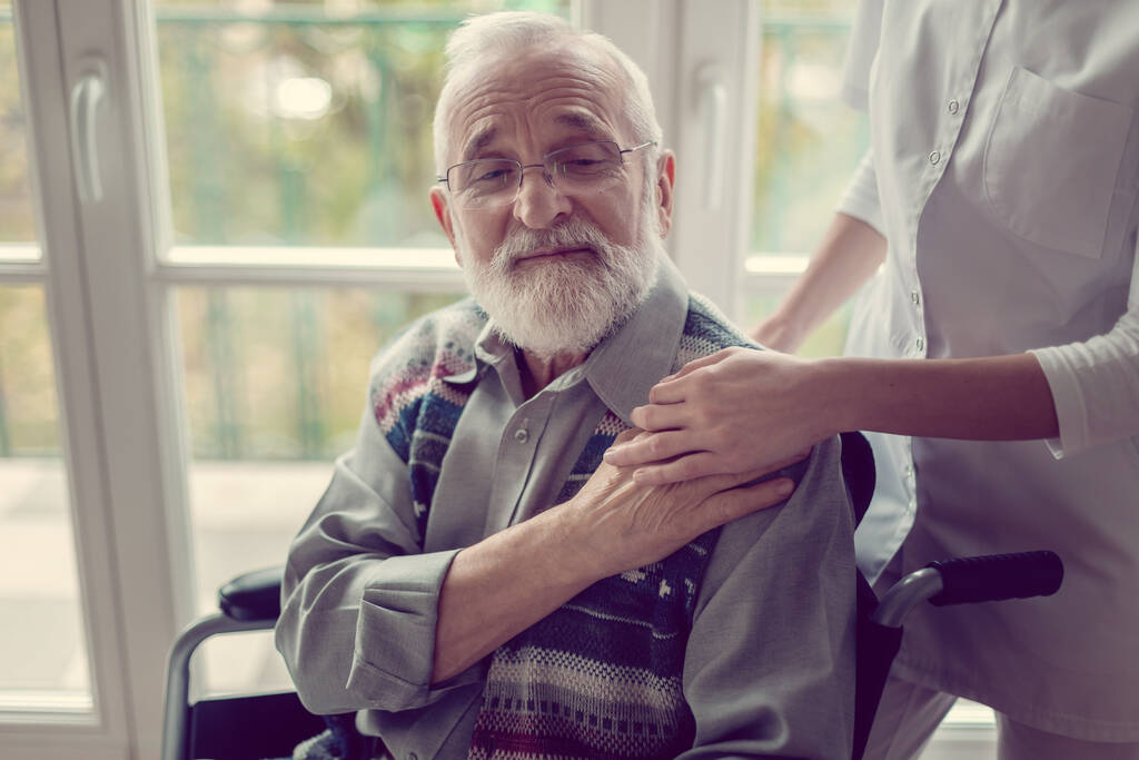 Homem mais velho doente sentado em uma cadeira de rodas e segurando a mão de sua enfermeira
 - Foto, Imagem
