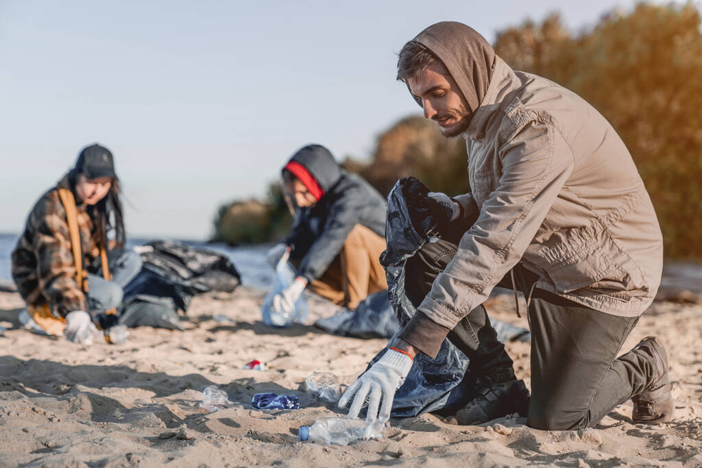 Bonito jovem voluntário com equipe reunindo lixo na praia
 - Foto, Imagem