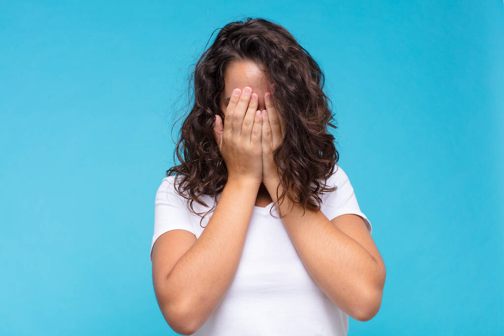 jonge mooie vrouw voelt zich verdrietig, gefrustreerd, nerveus en depressief, bedekken gezicht met beide handen, huilen tegen een blauwe achtergrond - Foto, afbeelding