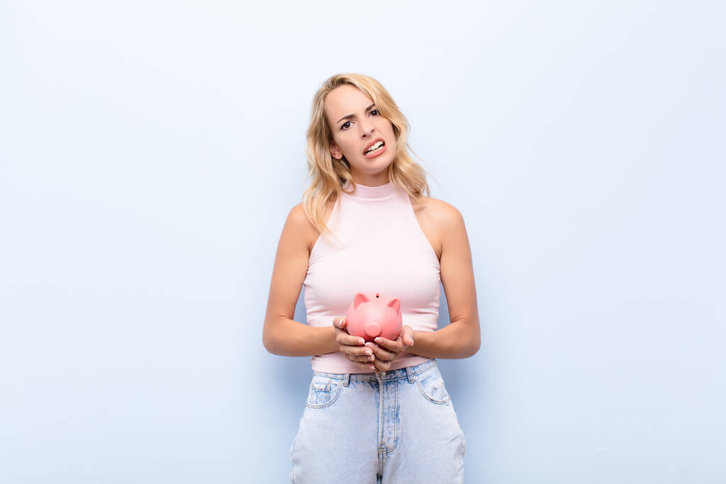 young blonde woman feeling puzzled and confused, with a dumb, stunned expression looking at something unexpected holding a piggybank - Foto, afbeelding