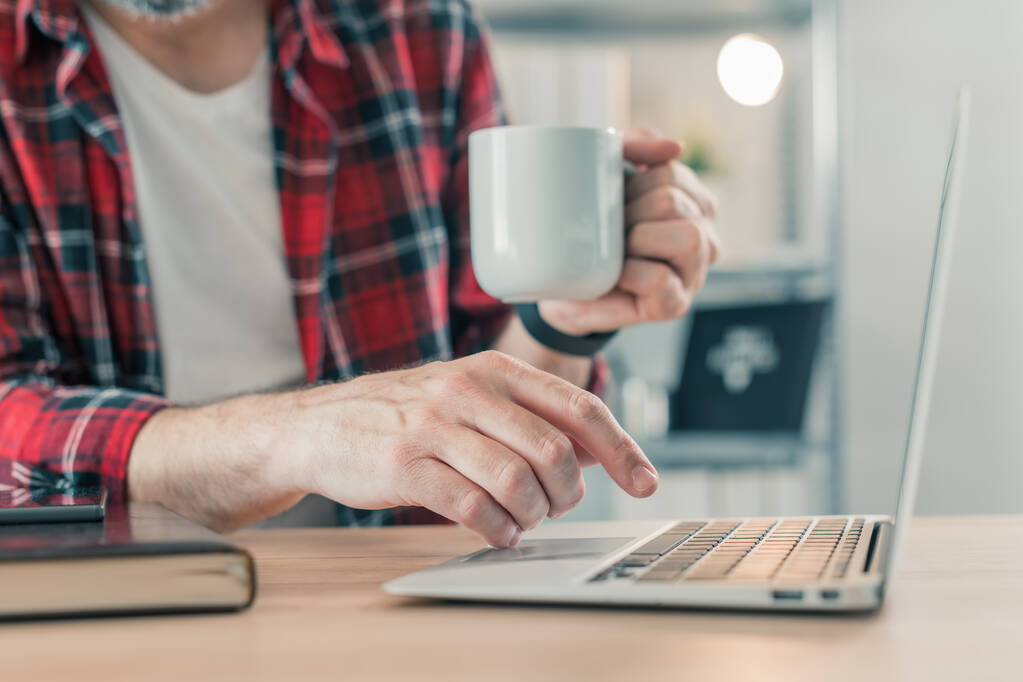 ノートパソコンや自宅のオフィスのインテリアでコーヒーを飲むフリーランサー、手に選択的な焦点 - 写真・画像