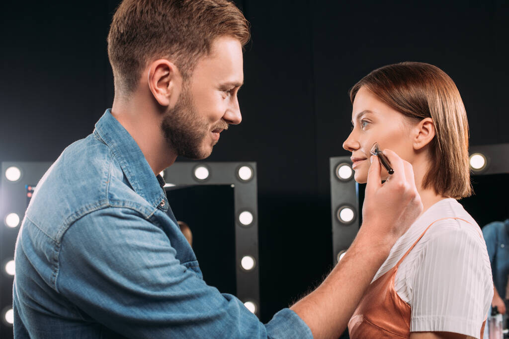 Seitenansicht des lächelnden Make-up-Artist hält kosmetische Pinsel in der Nähe Wange des Modells  - Foto, Bild
