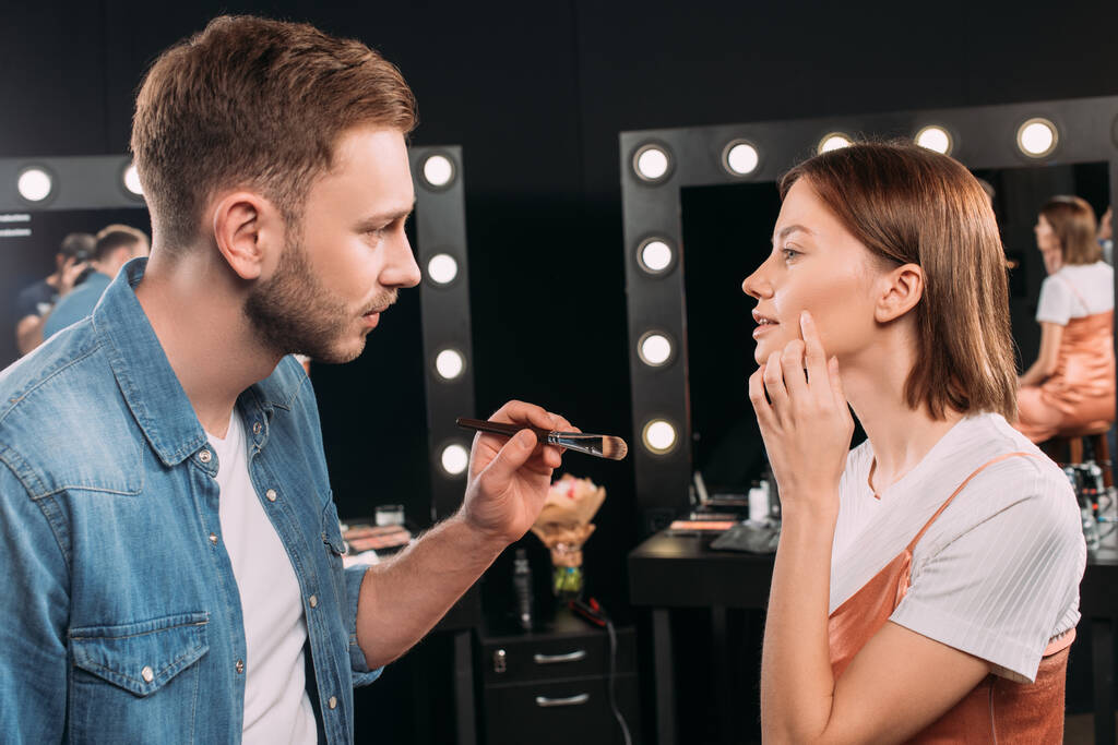 Vista lateral do modelo apontando na bochecha perto de maquiagem artista segurando escova cosmética no estúdio de fotos
  - Foto, Imagem