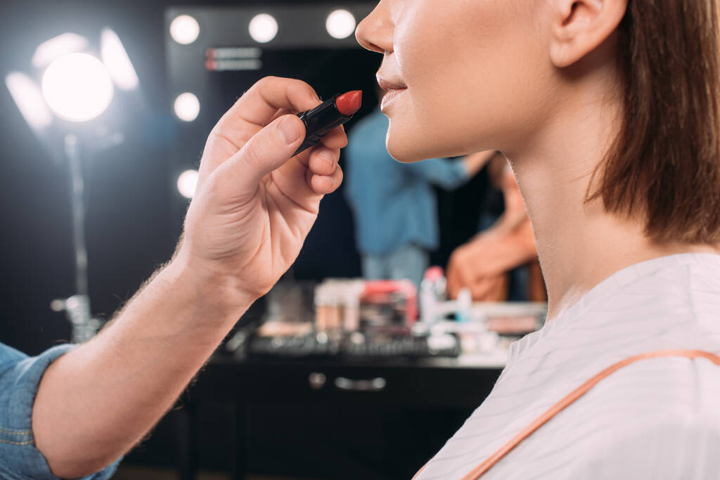 Vista recortada do artista de maquiagem segurando batom vermelho perto de modelo jovem no estúdio de fotos
  - Foto, Imagem