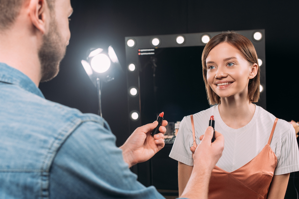Επιλεκτική εστίαση του καλλιτέχνη μακιγιάζ κρατώντας κραγιόν κοντά χαμογελαστό μοντέλο στο στούντιο φωτογραφιών  - Φωτογραφία, εικόνα