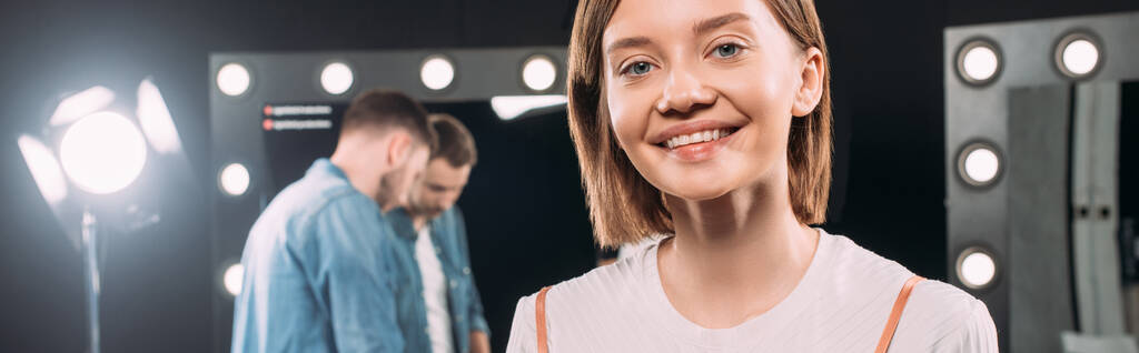 Panoramaausschnitt einer schönen lächelnden Frau, die in die Kamera blickt, während Make-up-Artist neben Spiegel im Fotostudio steht  - Foto, Bild