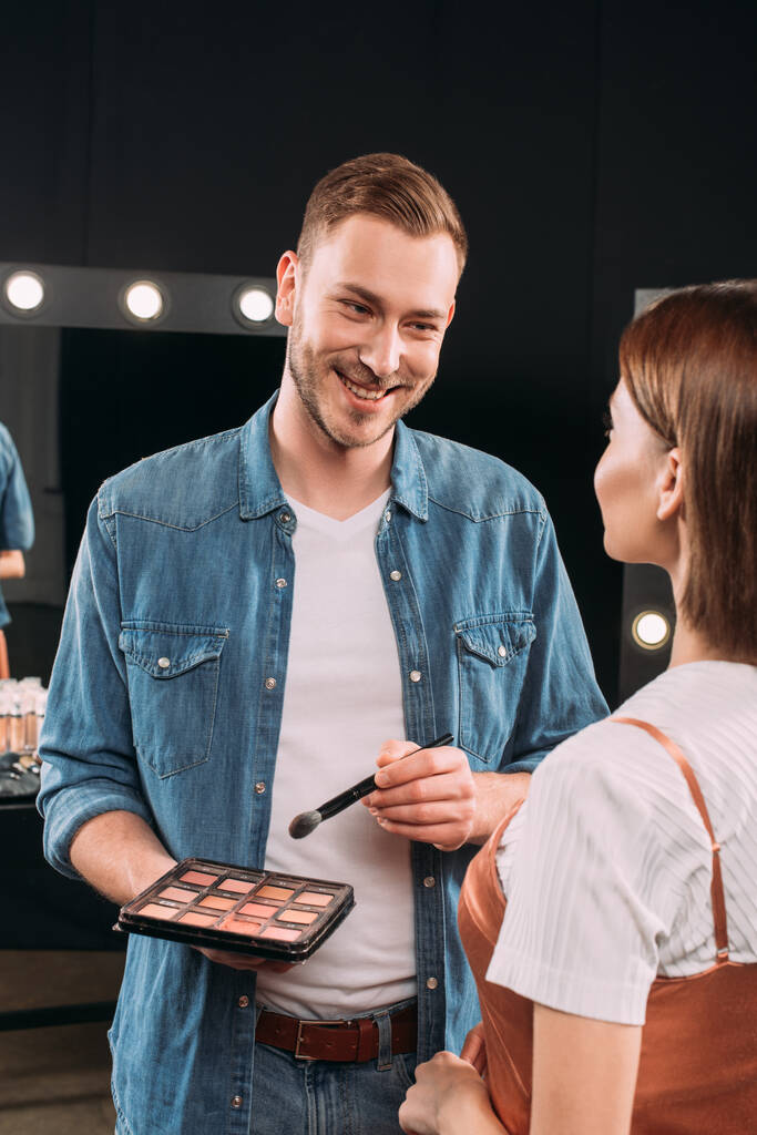 Selective focus of handsome makeup artist smiling at model while holding cosmetic brush and set of blush  - Photo, Image