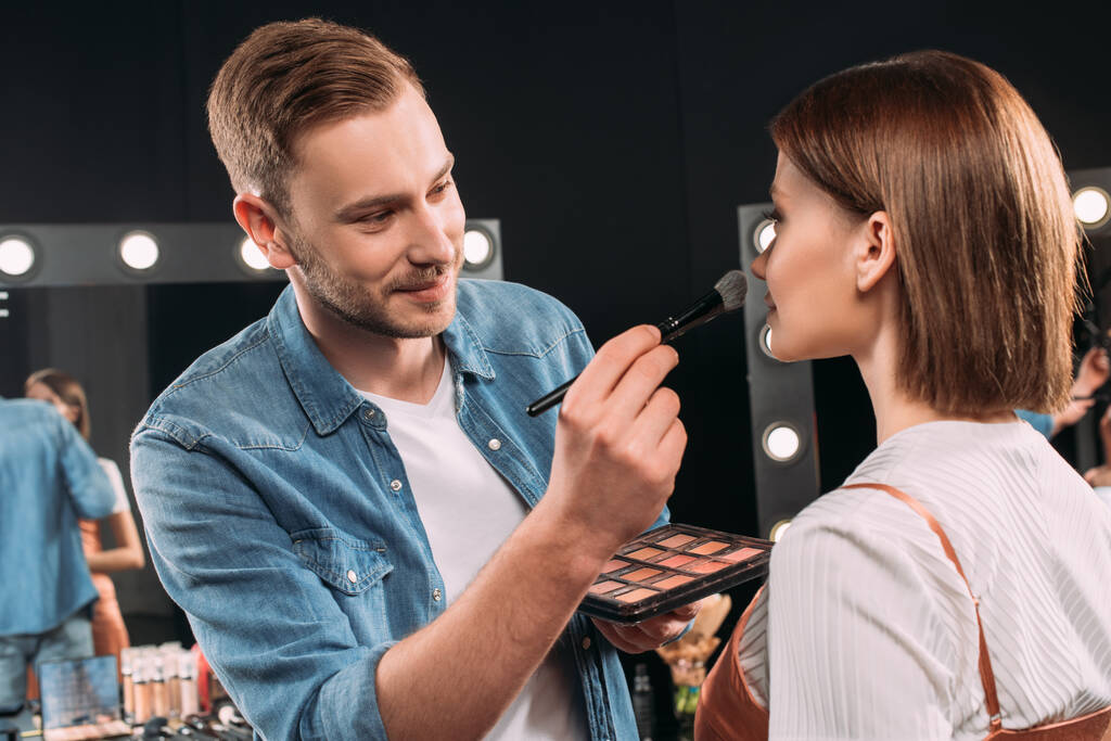 Sonriente artista de maquillaje aplicando rubor en la joven modelo en el estudio de fotos
  - Foto, Imagen