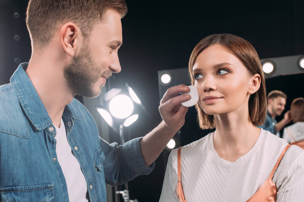 Usmívající se make-up umělec drží bavlněné podložky v blízkosti tváře krásného modelu ve fotostudiu  - Fotografie, Obrázek