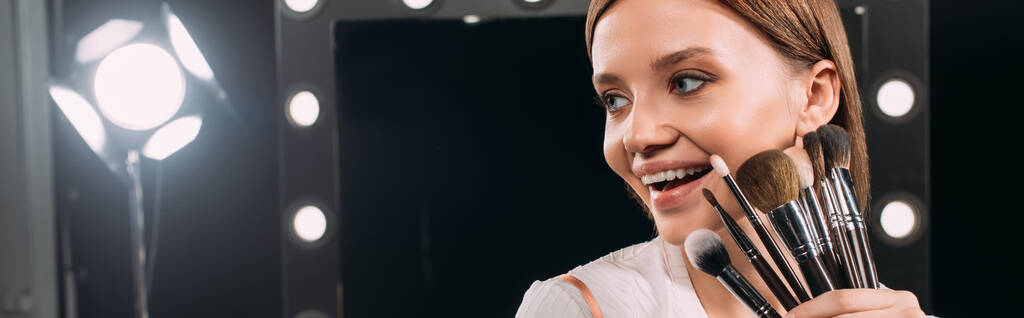 Панорамний знімок позитивної моделі, що тримає косметичні пензлі в фотостудії
  - Фото, зображення