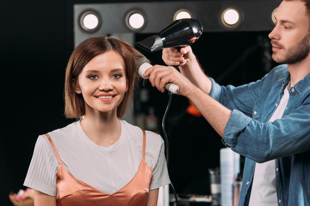 Knappe make-up kunstenaar doet kapsel met haarborstel en haardroger op glimlachend model in fotostudio  - Foto, afbeelding