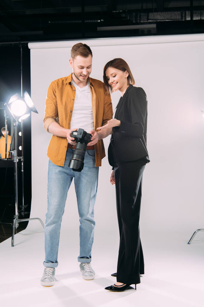 Piękny uśmiechnięty model wskazujący ręką na wyświetlacz aparatu cyfrowego w pobliżu przystojnego fotografa w studiu fotograficznym  - Zdjęcie, obraz
