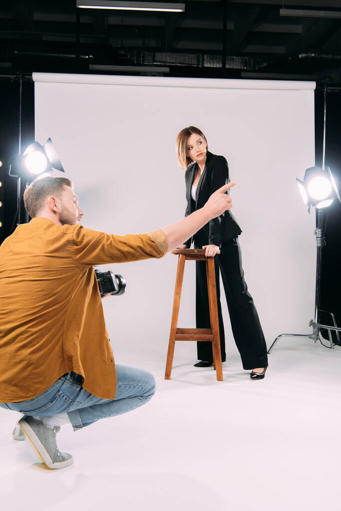 Fotógrafo señalando con el dedo a hermosa modelo cerca de la silla en el estudio de fotos
  - Foto, Imagen
