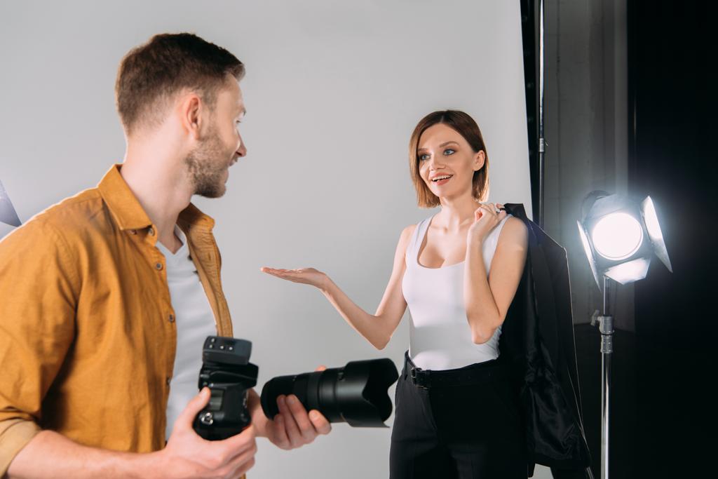 Улыбающаяся модель, держащая куртку и указывающая руками на фотографа с цифровой камерой в фотостудии
  - Фото, изображение