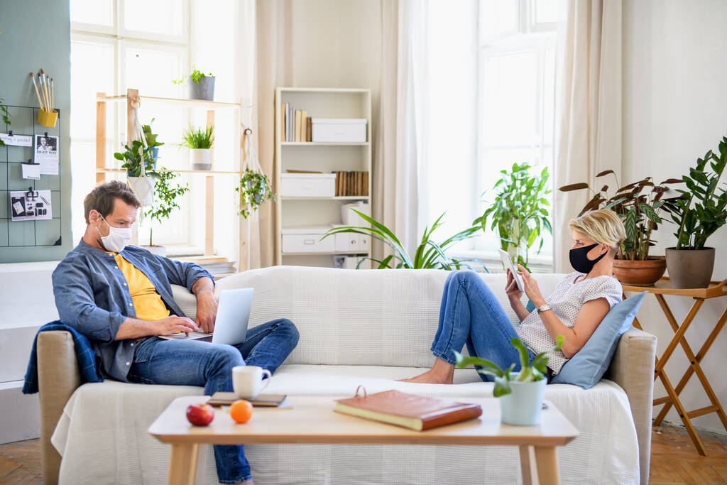 Ζευγάρι με μάσκες προσώπου κάθεται σε εσωτερικούς χώρους στο σπίτι, χρησιμοποιώντας τεχνολογία. - Φωτογραφία, εικόνα