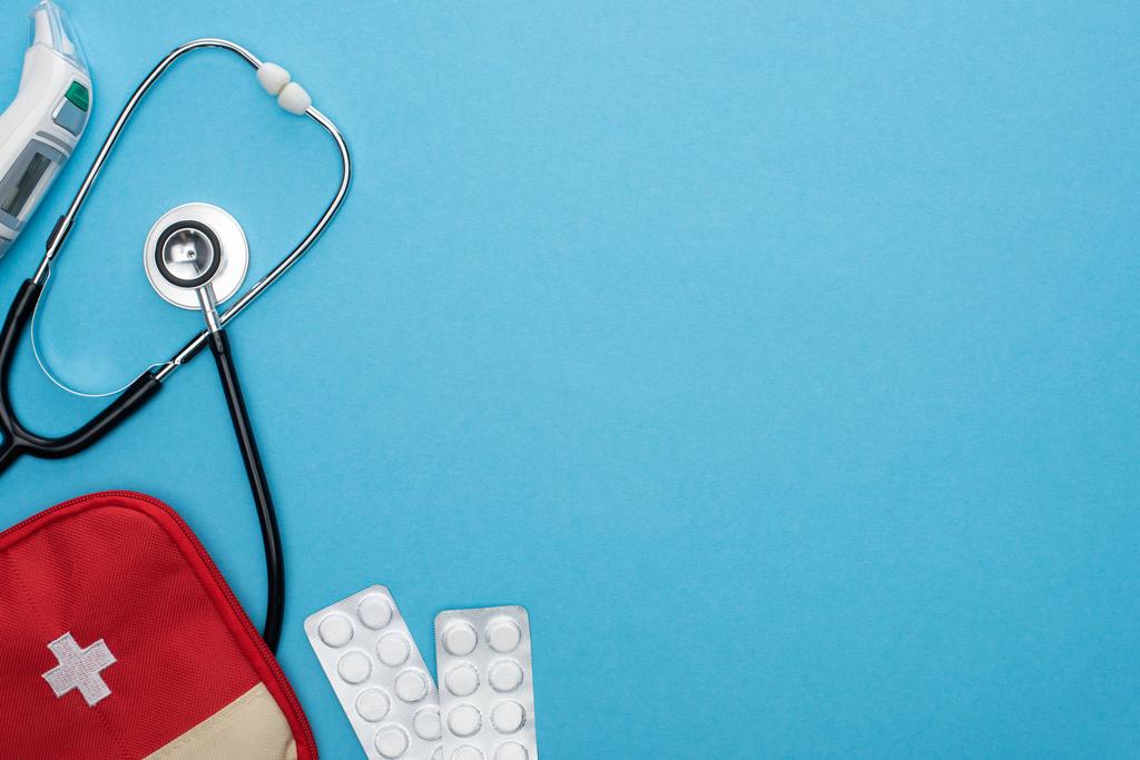 bovenaanzicht van pillen in blisterverpakkingen, verbanddoos, oorthermometer en stethoscoop op blauwe achtergrond - Foto, afbeelding