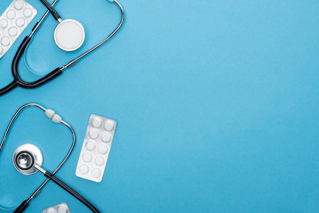 vrchní pohled na pilulky v blistrech a stetoskopy na modrém pozadí - Fotografie, Obrázek