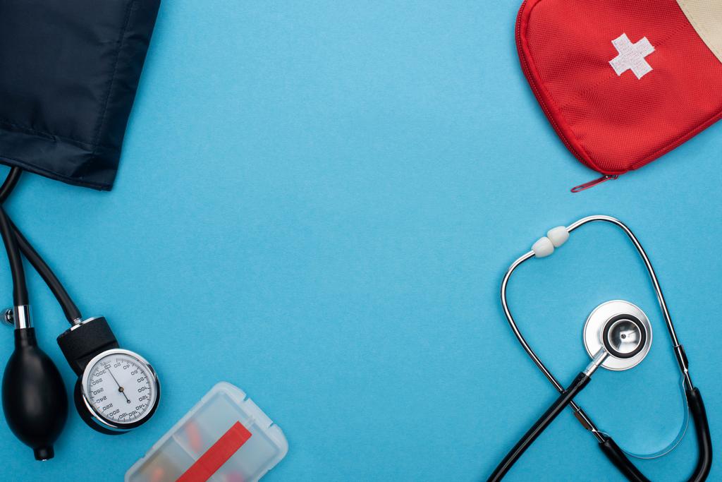 Ansicht von Pillen im Behälter, Blutdruckmessgerät, Verbandskasten und Stethoskop auf blauem Hintergrund - Foto, Bild