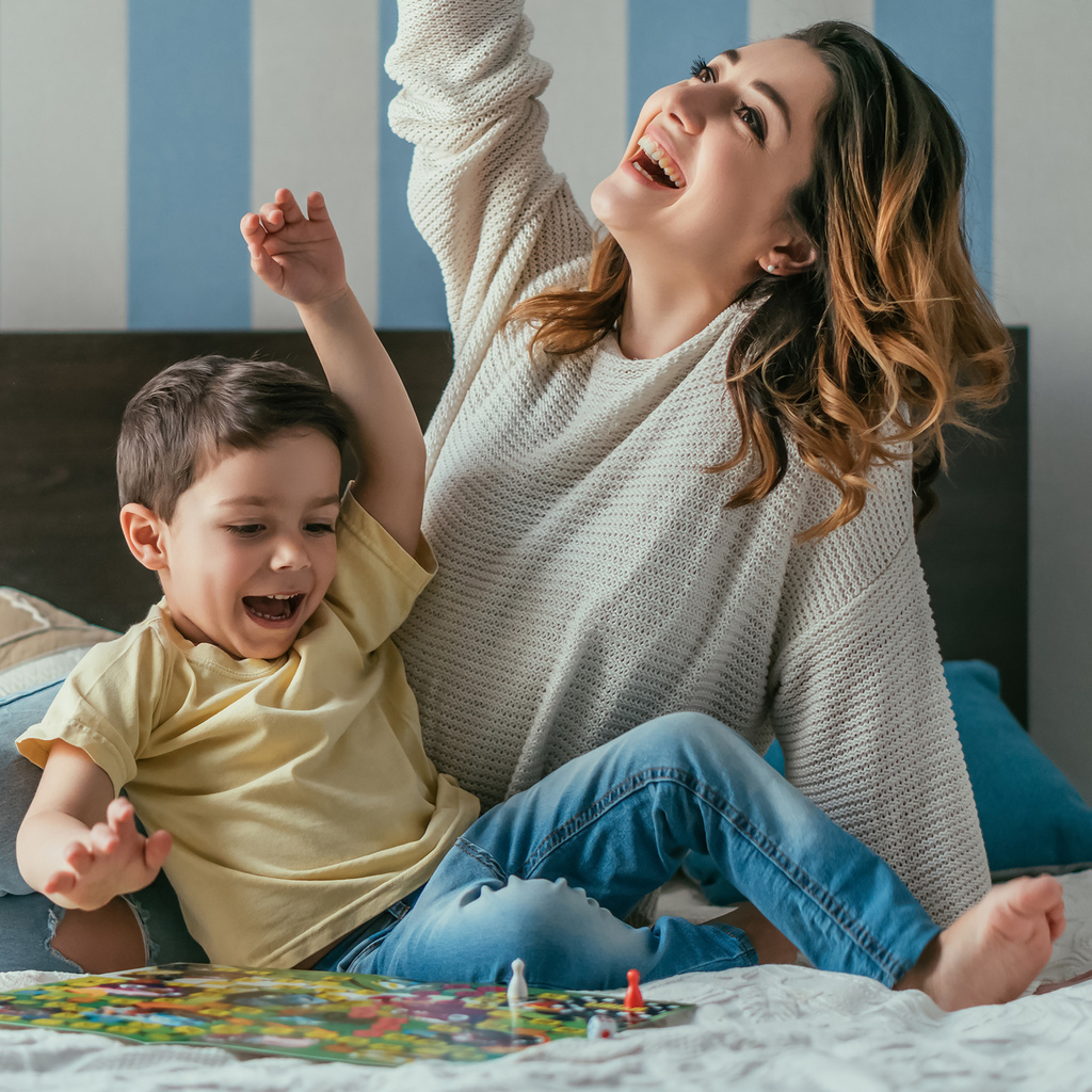 szczęśliwy, podekscytowany matka i syn pokazując gesty zwycięzcy podczas gry planszowej na łóżku - Zdjęcie, obraz
