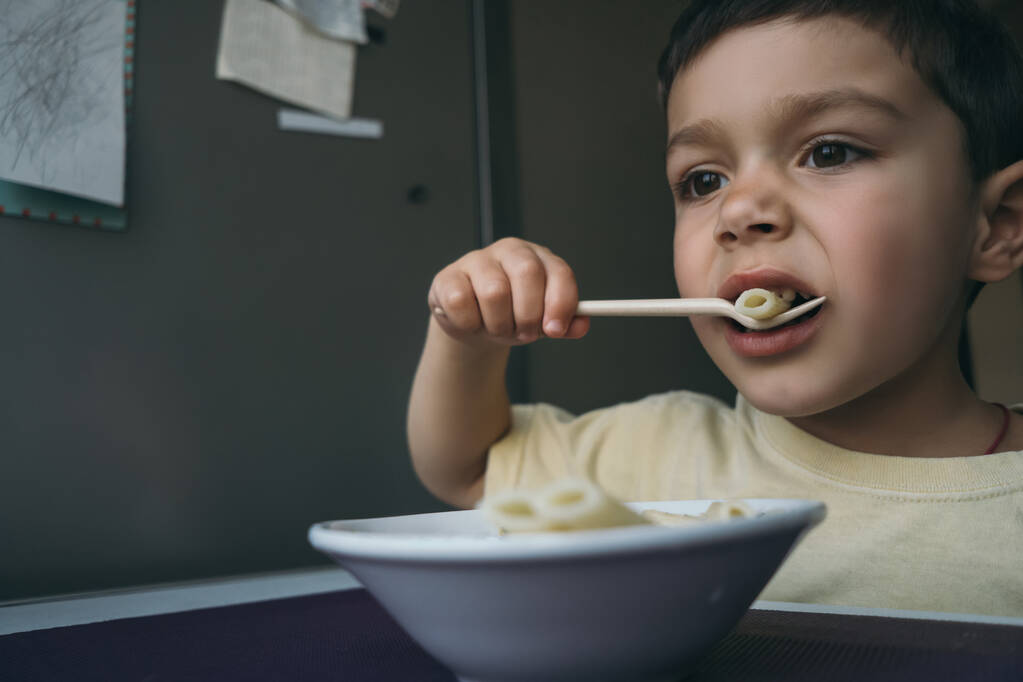 bonito morena menino comer penne pasta com garfo perto tigela na cozinha mesa
 - Foto, Imagem