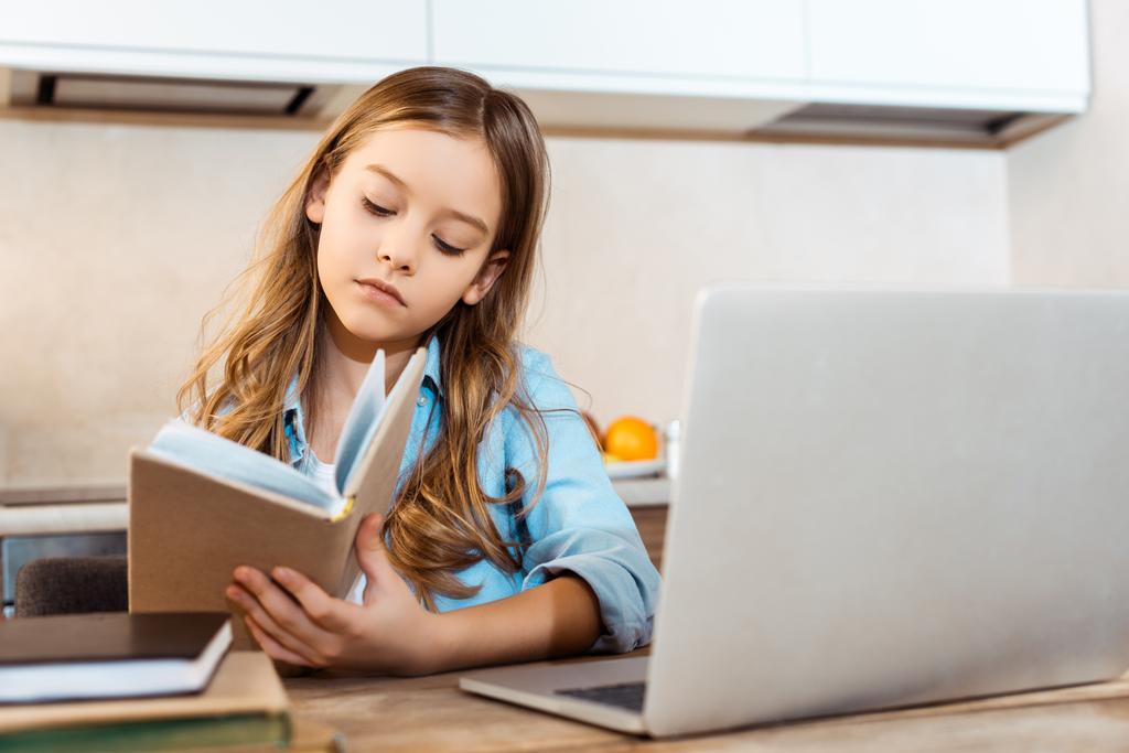 selective focus of cute kid holding book near laptop while online studying at home - Photo, Image