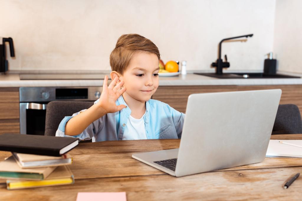 Evde e-öğrenim görürken el sallayan ve dizüstü bilgisayara bakan sevimli çocuğun seçici odağı - Fotoğraf, Görsel