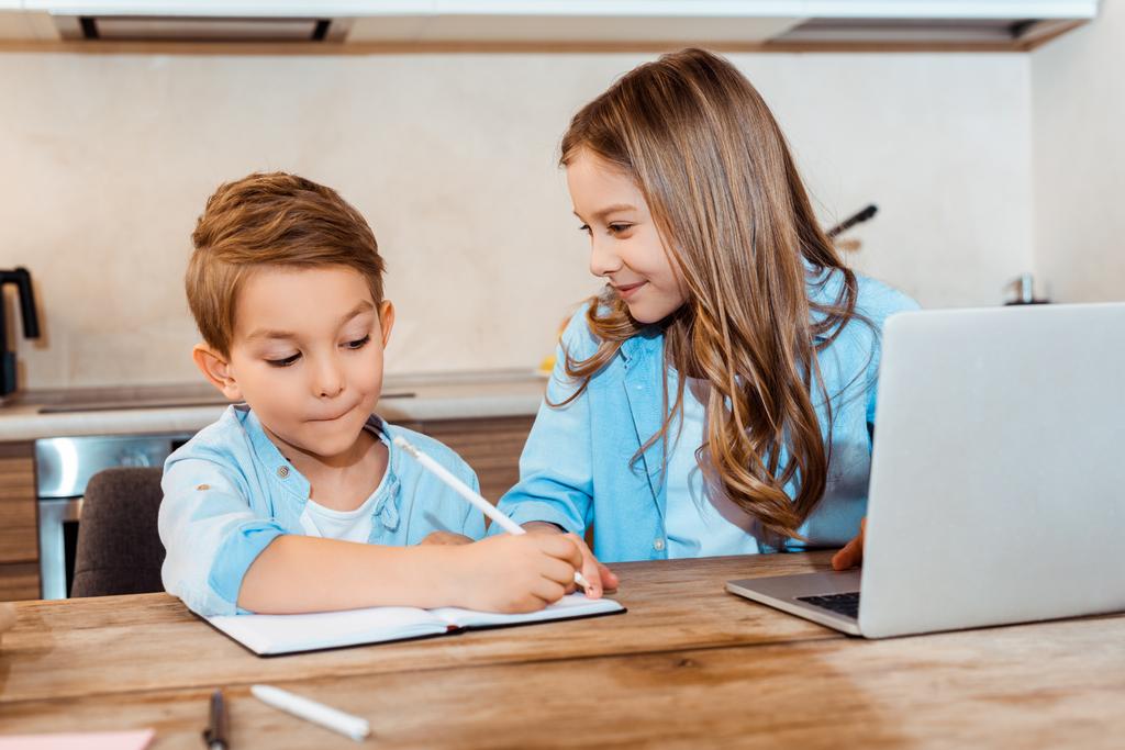 selektywne skupienie szczęśliwej siostry patrząc na brata piszącego w notebooku w pobliżu laptopa podczas e-learningu w domu - Zdjęcie, obraz