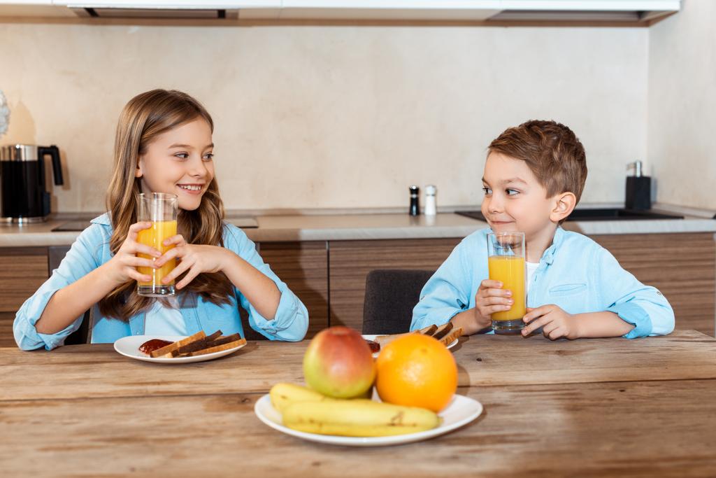 enfoque selectivo de hermanos felices mirándose unos a otros y sosteniendo vasos de jugo de naranja cerca del delicioso desayuno
  - Foto, Imagen