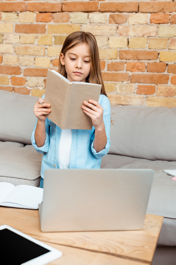 επιλεκτική εστίαση του χαριτωμένου βιβλίου ανάγνωσης παιδιών κοντά στο laptop και ψηφιακό δισκίο - Φωτογραφία, εικόνα