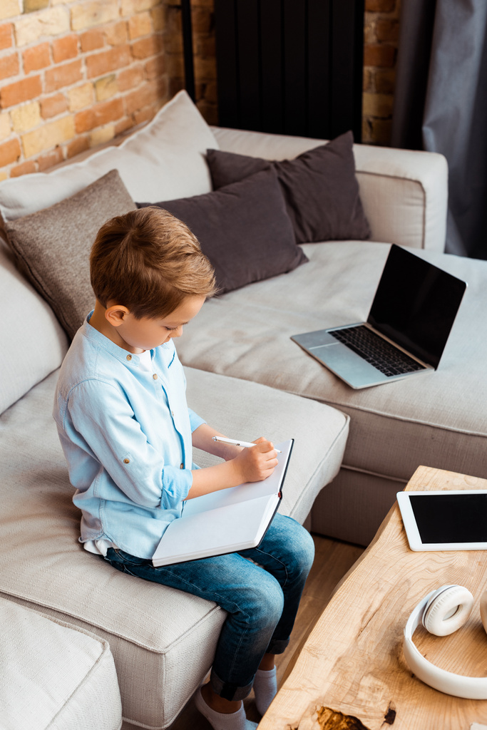 lindo chico escribiendo en cuaderno cerca de gadgets con pantalla en blanco mientras e-learning en casa
 - Foto, imagen