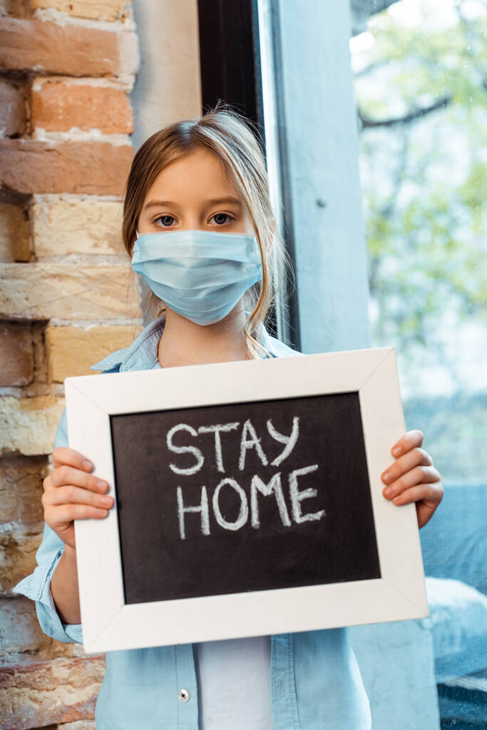 παιδί με ιατρική μάσκα κοιτάζοντας κάμερα και κρατώντας chalkboard με stay home lettering  - Φωτογραφία, εικόνα