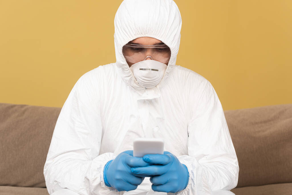Enfoque selectivo del hombre en traje de materiales peligrosos, guantes de látex y máscara médica utilizando el teléfono inteligente en el sofá en casa
  - Foto, imagen
