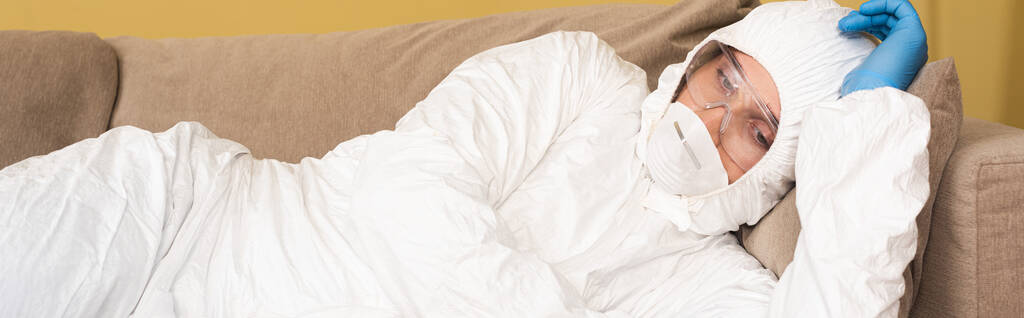 Panoraama suunta mies lääketieteellinen naamio, suojalasit ja hazmat puku makaa sohvalla
  - Valokuva, kuva