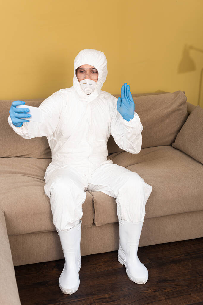 Muž v hazmat obleku a lékařské masky mávání rukou při braní selfie s smartphonem na gauči doma  - Fotografie, Obrázek