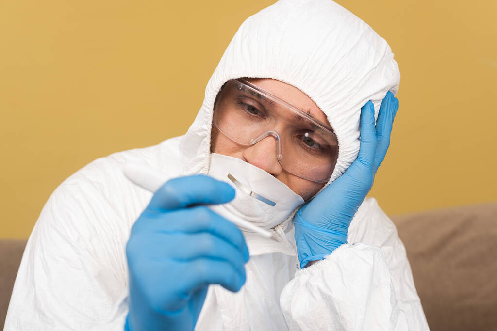 Selective focus of upset man in hazmat suit and latex gloves holding thermometer at home  - Photo, Image
