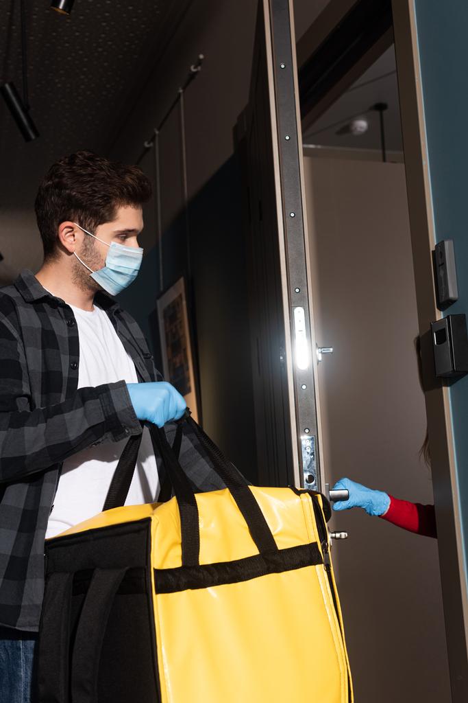 Vue latérale du courrier dans un masque médical tenant un sac thermique près de la femme dans la porte d'ouverture du gant en latex
  - Photo, image