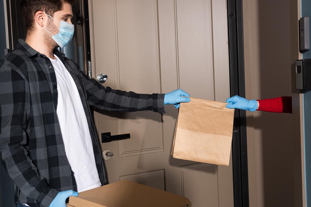Oldalnézet a szállító férfi ad csomagot a nő latex kesztyű közelében nyitott ajtó  - Fotó, kép