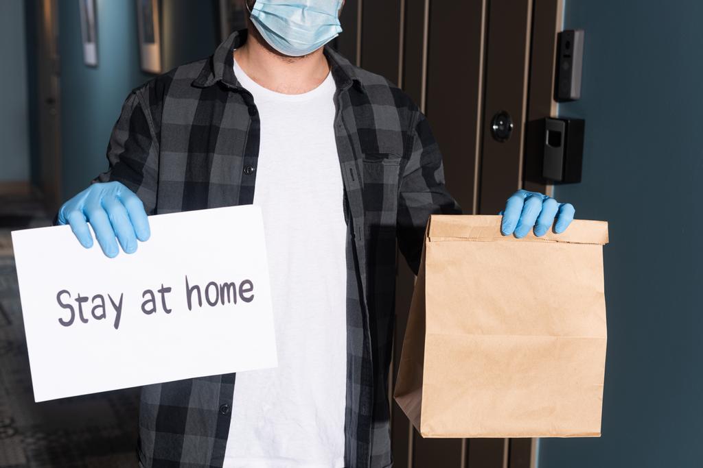 Cropped view of courier in medical mask holding package and card with stay at home lettering on entryway - Photo, Image