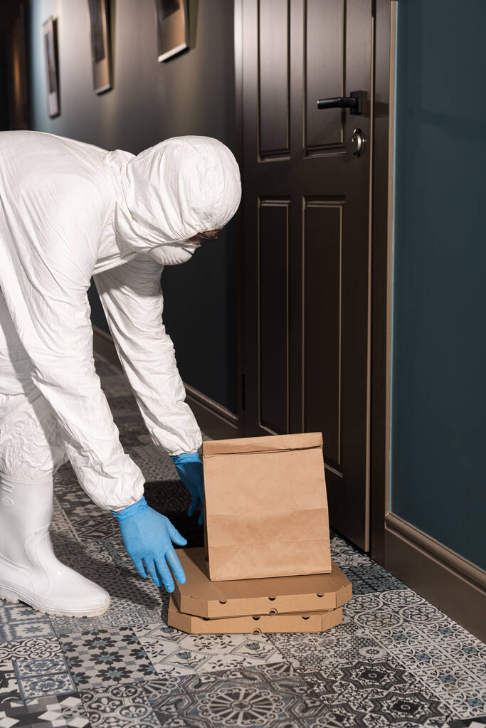Zijaanzicht van de levering man in hazmat pak en medisch masker zetten pakket en pizza dozen op de vloer in de buurt van de deur op de veranda - Foto, afbeelding