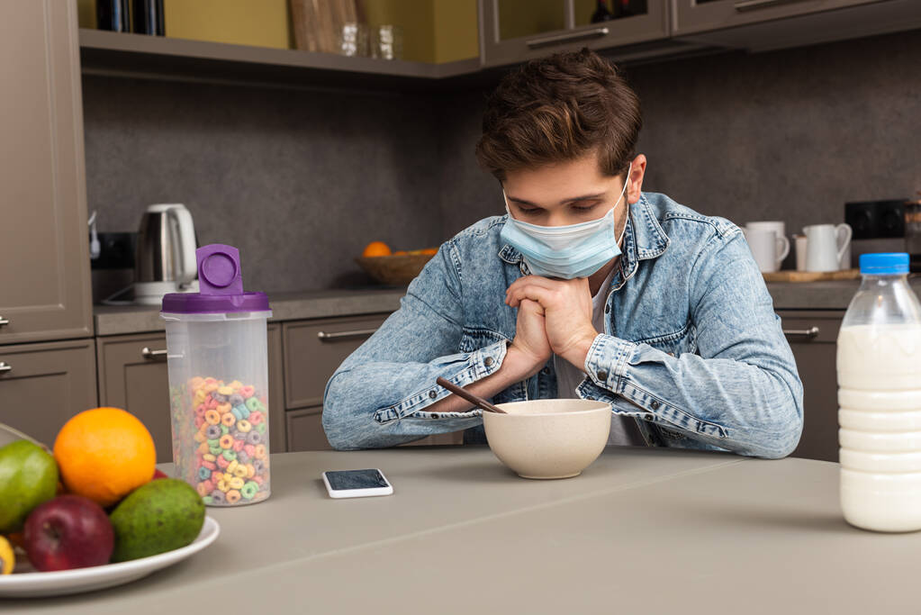 Concentration sélective de l'homme dans le masque médical assis près des céréales, bouteille de lait et de fruits dans la cuisine
  - Photo, image