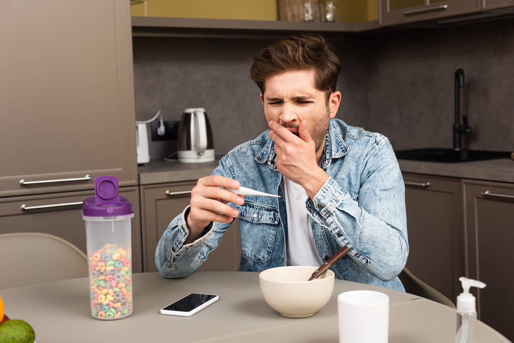 Человек зевает, держа термометр рядом с зерновыми и смартфоном на столе
  - Фото, изображение