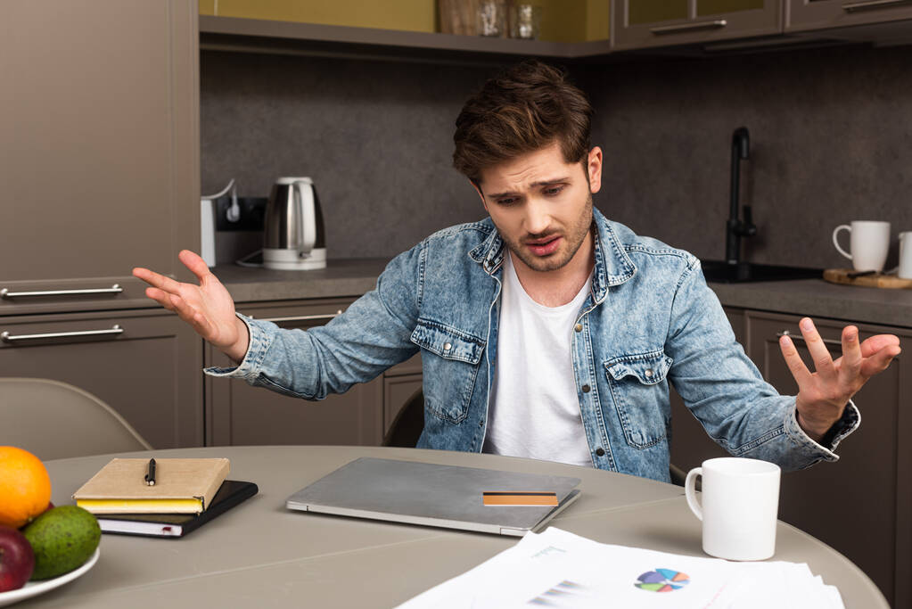 Concentration sélective de l'homme inquiet regardant la carte de crédit sur l'ordinateur portable près des papiers sur la table de cuisine
  - Photo, image